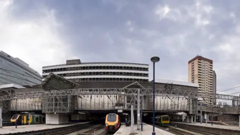 An exterior view of Birmingham New Street station showing multiple tracks and platforms side by side. There are three trains in view on the tracks, with a high rise building to the right in the background