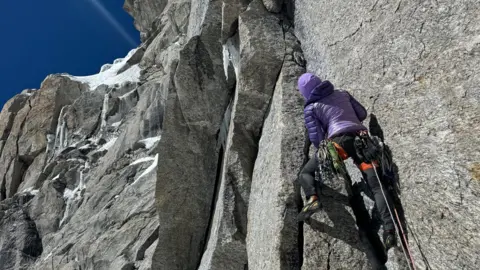 Fay Manners Una mujer vestida de púrpura, de espaldas a la cámara, sube la ladera de una gran montaña gris con un cielo azul en la distancia.