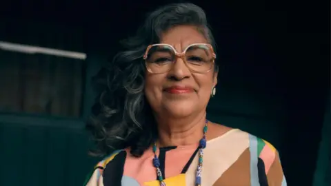 A woman with long dark hair, large glasses and a colourful patterned top in front of a dark background