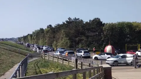 RNLI Cars parked along a single carriageway road, stretching into the distance. There are trees on the right of the road, and a grassed, sloping, sea wall bank on the left. There is a large metal ball, painted red and yellow with cars on either side. A metal gate blocks access to a paved area in the foreground.
