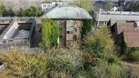 A tall dilapidated old building, with frost on its grey tiled roof. It has broken window panes and a wooden structure that juts out that is broken. It is surrounded by industrial buildings and structures as well as overgrowth.