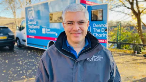 BBC/Mark Ansell Andy Evans standing outdoors in front of a large blue mobile unit. The unit has text on its side that reads “Dentaid The Dental Charity” along with a logo featuring a stylised tooth. There is also additional text mentioning “Bupa Foundation” and a contact number partially visible. He is wearing a dark jacket with the same “Dentaid The Dental Charity” logo printed on the left chest area, and underneath the jacket, a blue collared shirt is visible.