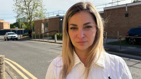 Martin Heath/BBC Joanna Wade with long blonde hair, looking at the camera and wearing a white shirt. There is a single-storey brick court building behind her, with a railing around the roof. There are concrete bollards along the pavement outside, and a narrow road behind Wade, with double yellow lines.