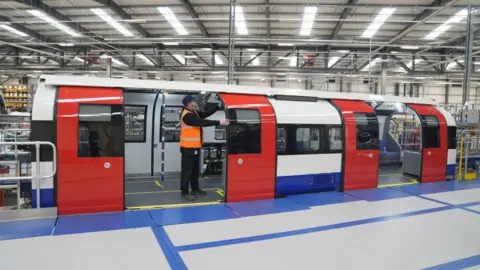 A blue, red and white train carriage inside a factory. The two sets of red doors of the carriage are open. A man wearing an orange hi-vis vest is stood in the doorway of the left set of doors, with his hands holding on to the side of one of the doors.