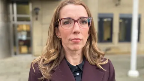 BBC/Emma Glasbey A woman with shoulder length brown hair and large spectacles stands in front of a blurred court building