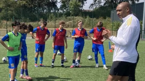 Six teenage boys in blue football kits, wearing red bibs, stand on a football pitch looking at a football coach who is dressed in white and giving them instructions. There are white and black footballs at their feet.