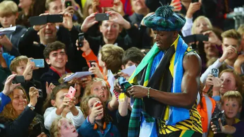 Getty Images Usian Bolt signs autographs for fans waiting trackside during the Commonwealth Games in 2014. Bolt is wearing a tartan hat, and has a Scottish flag and tartan scarf wrapped around his shoulders. He is wearing his running gear in yellow and green colours. The fans are all smiling and reaching out with things for him to sign. 