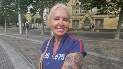 Aisha Iqbal/BBC A woman in a purple zip-up t-shirt with white-blond hair and tattoos on her arm. She smiles at the camera.