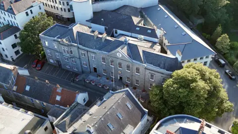 An aerial view of the States of Guernsey building. It is a large grey stone building, surrounded by streets and trees.