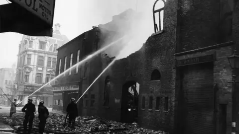 Getty Images A black and white photo of several firefighters aiming jets of water at a bomb-damaged, blackened building, which has lost its roof and has jagged walls and blown-out windows. The street is covered in rubble.
