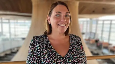 The image shows Heledd Fychan standing in the Senedd looking directly at the camera and smiling. She is wearing a colourful dress. The Senedd's funnel is behind her. It is a head and shoulders shot of her.