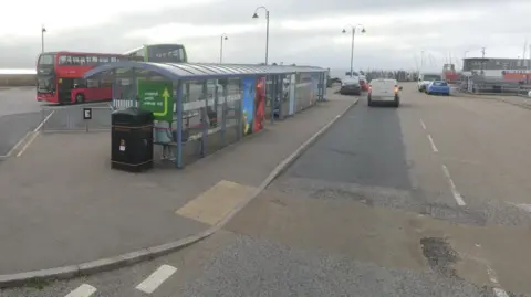 Two double decker buses - one red and one green - are in the background behind a covered bus bay. There is a junction to the left and a road to the right which leads to a car park.