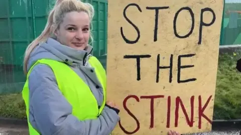 A young woman with long blonde hair, in a grey coat and yellow high vis vest, holds a sign that says 'stop the stink' in front of large green skips at a landfill site.