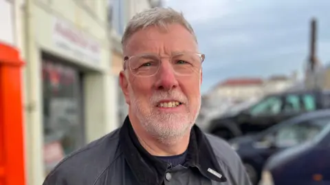 Deputy Adrian Gabriel from the States of Guernsey stood outside some shops and parked cars. He is looking straight at the camera and is smiling. 