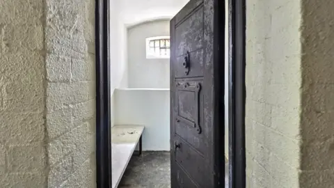 An imposing heavy grey metal cell door stands ajar, a bed can just be seen inside.