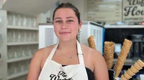  Mieke is stood in front of ice cream cones and an ice cream machine in the shop. She is wearing an apron and has short brown hair.