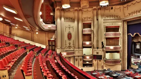 The ornate interior of Streatham Hill Theatre, featuring rows of red seats, decorative balconies, tall columns and period architectural detailing surrounding the auditorium.