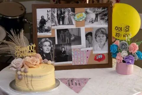 Barrowhill Hall A montage of black and white photos of Jean, next to a 100th birthday cake with flowers on it and a "Happy 100th Birthday" topper, as well as a yellow balloon and flowers