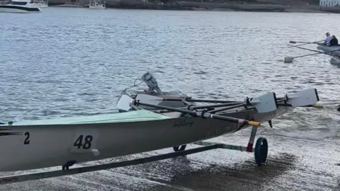 BBC A swift style rowing boat on the slip way above the water. It is long with a number of ores inside it. There are some rowers in another boat in the background along with some larger boats on the water. 