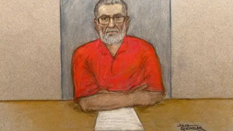 Julia Quenzler/BBC Court sketch showing a bearded Peter Sullivan, wearing a red open-necked shirt and glasses, sitting at a desk with some papers in front of him.