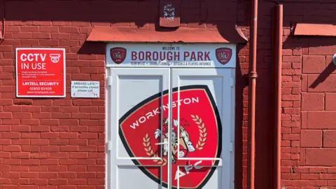 The players' entrance at Borough Park in Workington. It is a white double door with the red badge of Workington AFC and across it. A sign above reads Welcome the Borough Park and lists those allowed to enter. It is set into the outer brick wall of the ground which has been painted a brownish red. A red sign warns of CCTV in use and sits next to a smaller white sign which gives a phone number for latecomers who have arrived after the turnstiles have closed.