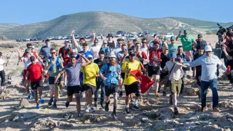 PA Media Ras is surrounded by riding cook supporters as he crosses a finish line in Tunisia. He is wearing a blue England football shirt and a bucket hat. 