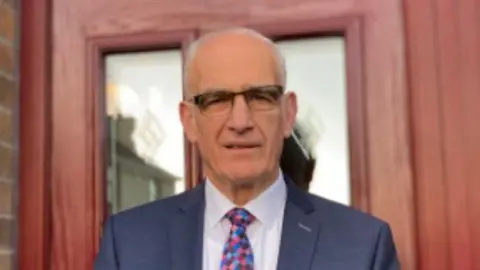 The Reverend John Greer The Rev John Greer - a bald man looking to camera and wearing glasses, a dark blue blazer, white shirt and multi-coloured tie