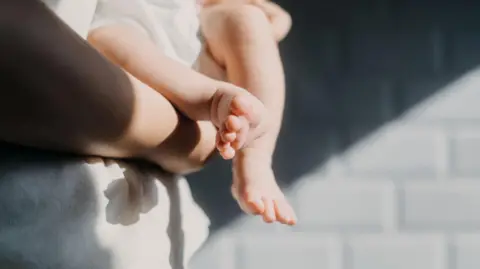 Getty Images Close-up of a baby's feet. The baby is held by an adult wearing a white shirt, standing in front of a grey brick wall