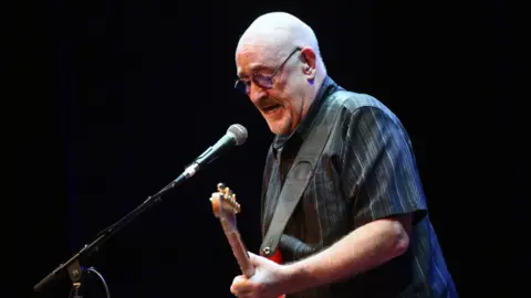 Scott Dudelson/Getty Images Dave Mason in a dark striped shirt with a red guitar strapped over his shoulder. He is playing the guitar and next to a microphone in front of a black background.

