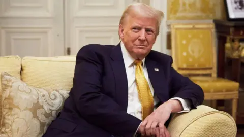 Donald Trump, an elderly man with blonde hair, sits on a golden couch. He is wearing a blue suit with a white shirt and gold tie