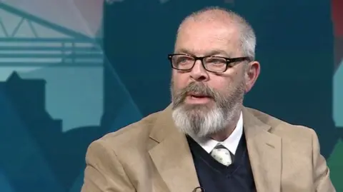 Councillor Stephen Place sitting in the Politics North studio during a live broadcast. He is wearing a light brown jacket over a dark jumper and white shirt and tie.
