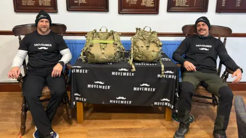 Nathan Curtis and Matthew Clarke lean back as they sit in carver chairs either side of a table where their camouflage vests are placed side-by-side. They are both wearing black winter running gear and beanie hats with Movember t-shirts bearing the white moustache logo. The table is draped with a black flag with the Movember logo repeated across it.