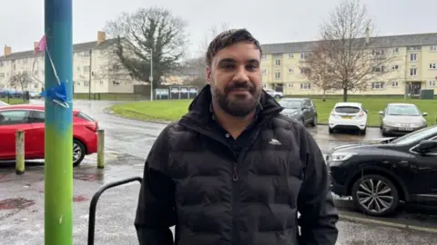 A photograph of independent councillor Alastair Chambers. He's wearing a black coat and standing in front of a road, a green and some homes. It's raining and there's a few trees with no leaves on the branches. 