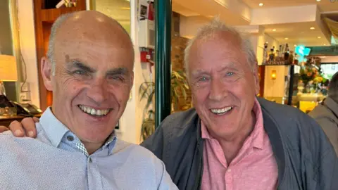 Tony Jones Jones, wearing a blue shirt, sitting next to Martin Tyler, who is wearing a grey jacket over a pink polo shirt. Both are smiling and inside a restaurant.