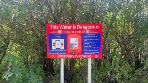 A read board that read: "This water is dangerous. Do not Enter!" It contains an emergency throwline and instructions on how to use it.