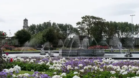 Friends of Stanley Park General view of the park with purple and white flowers in the foreground in front of a fountain. There are trees in the background with the clock tower peeking over the top of them to the left