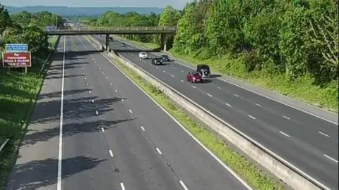 A stretch of the M5 through rural Somerset on a sunny day. The carriageway on the left is completely empty, with no traffic travelling down it, while the carriageway on the right has about ten cars travelling up. There is a bridge over the motorway and brown signs at the roadside.