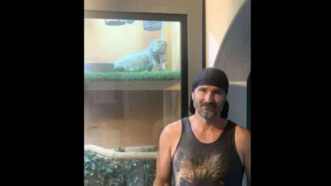 Dean Tyrrell A man in a dark grey singlet and dark grey bandana is standing in front of a vivarium. A blue-coloured iguana is inside the enclosure.