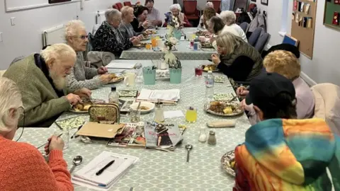 A large group of older people are sat round two tables, with green and while table cloths, eating lunch and socialising at their weekly lunch club.