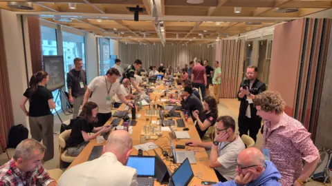 MEWC UK UK championship competitors sat around a very long table in a room full of people. The scene looks very buzzy and lots of people are sat in front of computers.
