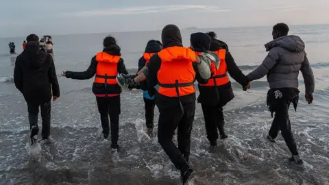 Getty Images লাইফ জ্যাকেট পরে পানির ধারে হাঁটছেন একজন শিশুসহ বেশ কয়েকজন