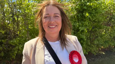 A woman with brown hair is wearing a cream coloured jacket with a red Labour Party rosette. She is smiling at the camera and standing in front of a green hedge.