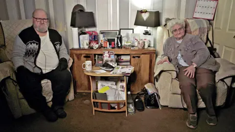 Paul and Christine sat in the armchair in their living room. Paul is on the left hand side, with brown hair, glasses and beard. He is wearing a white T-shirt, black trousers, shoes and a pattern zipped cardigan. He is wearing black woolen gloves. Christine has short brown hair and glasses. She wears a lilac cardigan with applications flowers, trousers and shoes. They have lamps and family photos with a wooden cabinet, and a small table with coffee mugs. 