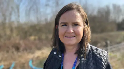 Tom Jackson/BBC Collette O'Shea has shoulder-length brown hair and is wearing a blue lanyard and a black coat. She is standing next to a wired fence which is in front of a row of trees.