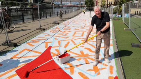 Sir Brendan Foster, an older man wearing a black polo shirt and beige trousers, using a paint roller with a very long handle to pose as if painting a red section of the mural. The artwork, which is still being created, is currently predominantly white with different orange shapes marked seemingly randomly along its length but perhaps as guide marks for the other elements of the design. It sits on the block-paved quayside path with the River Tyne to the left. It sits on the Newcastle bank of the river close to the Tyne Bridge. In the distance the Swing Bridge, High Level Bridge and blues steel QE2 Bridge can be seen on what is a very sunny day. There are still temporary silver metal fences around the mural which is also being worked on by a man in a hi-vis waistcoat in the background. A couple of cyclists are travelling past in opposite directions.