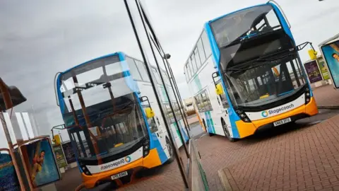An empty blue, white and orange Stagecoach branded double decker bus next to a bus stop. Its reflection can also be seen in the windows of another bus parked next to it, only part of which is in shot.