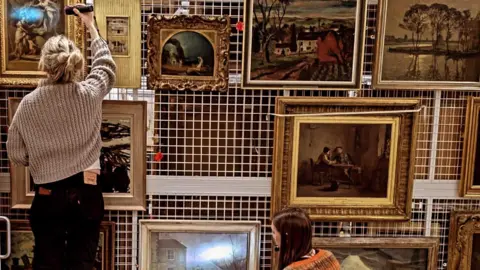 Critchlow & Kukkonen Ltd Two women use torches to shine lights on a variety of portraits, arranged on a white rack. 