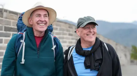 Brian Mole (left) and Melvyn Mole (right) at the Great Wall of China. Brian is wearing a Panama hat, red T-shirt, green fleece sweater and has a rucksack on his back. Melvyn is wearing a khaki baseball cap, blue T-shirt, a black zipped top and has a black scarf around his neck. He has a while and black bag strap over his right shoulder.
