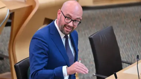Getty Images Ben Macpherson speaking in the Scottish Parliament, while gesturing with his right hand. He is a bald man with glasses and a beard and is wearing a blue suit, white shirt and a tie.
