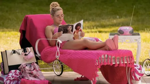 A woman in a pink bikini lies on a deck chair covered in pink blankets, reads a magazine. there are pink towels, a tote bag and a radio next to her.